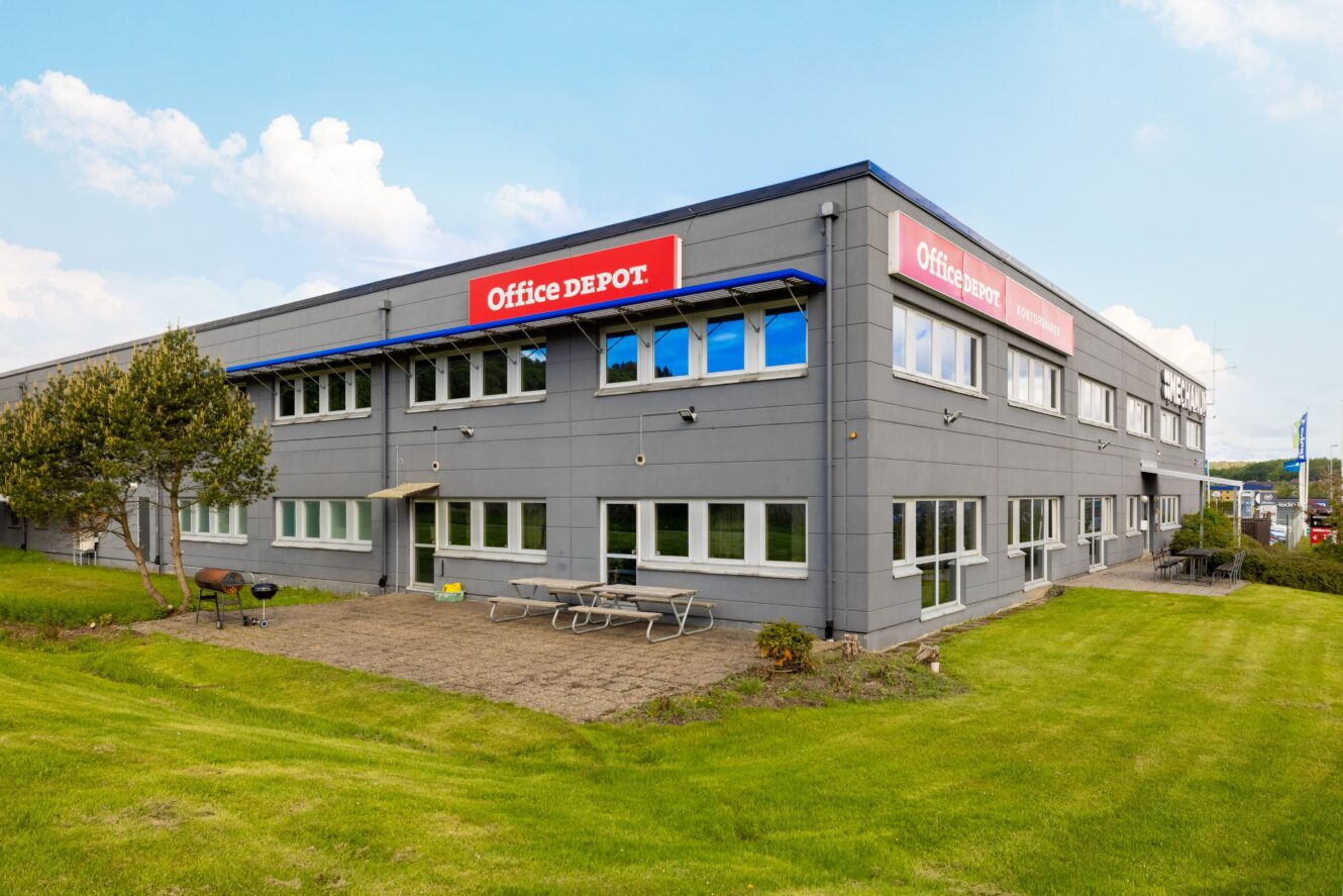 En modern grå byggnad med Office Depot-skyltar, omgiven av gräs, träd och picknickbord utomhus under en delvis molnig himmel.