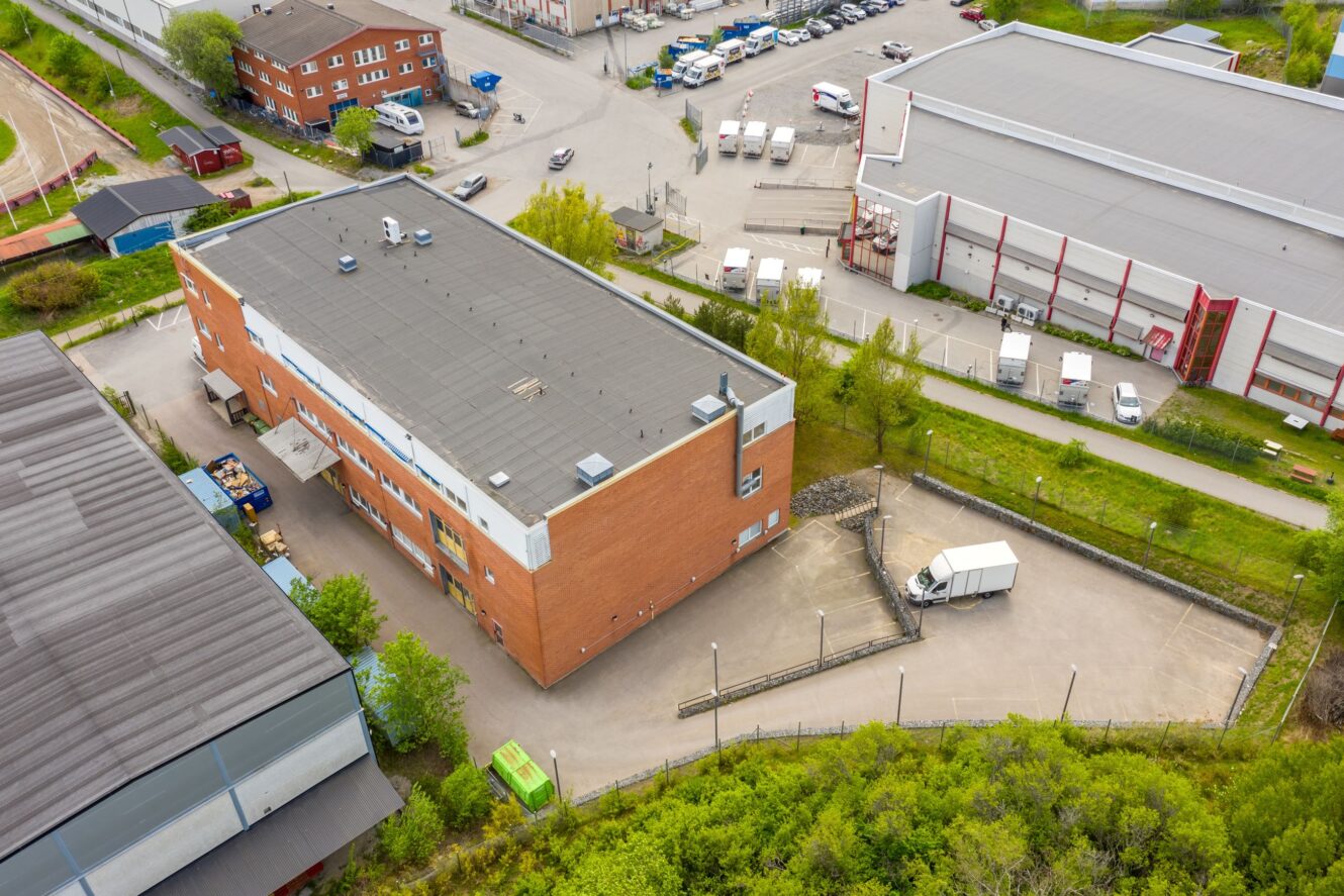 Flygfoto över en kommersiell tegelbyggnad med en lastbil parkerad vid en lastkaj, omgiven av andra industribyggnader, vägar och grönska.