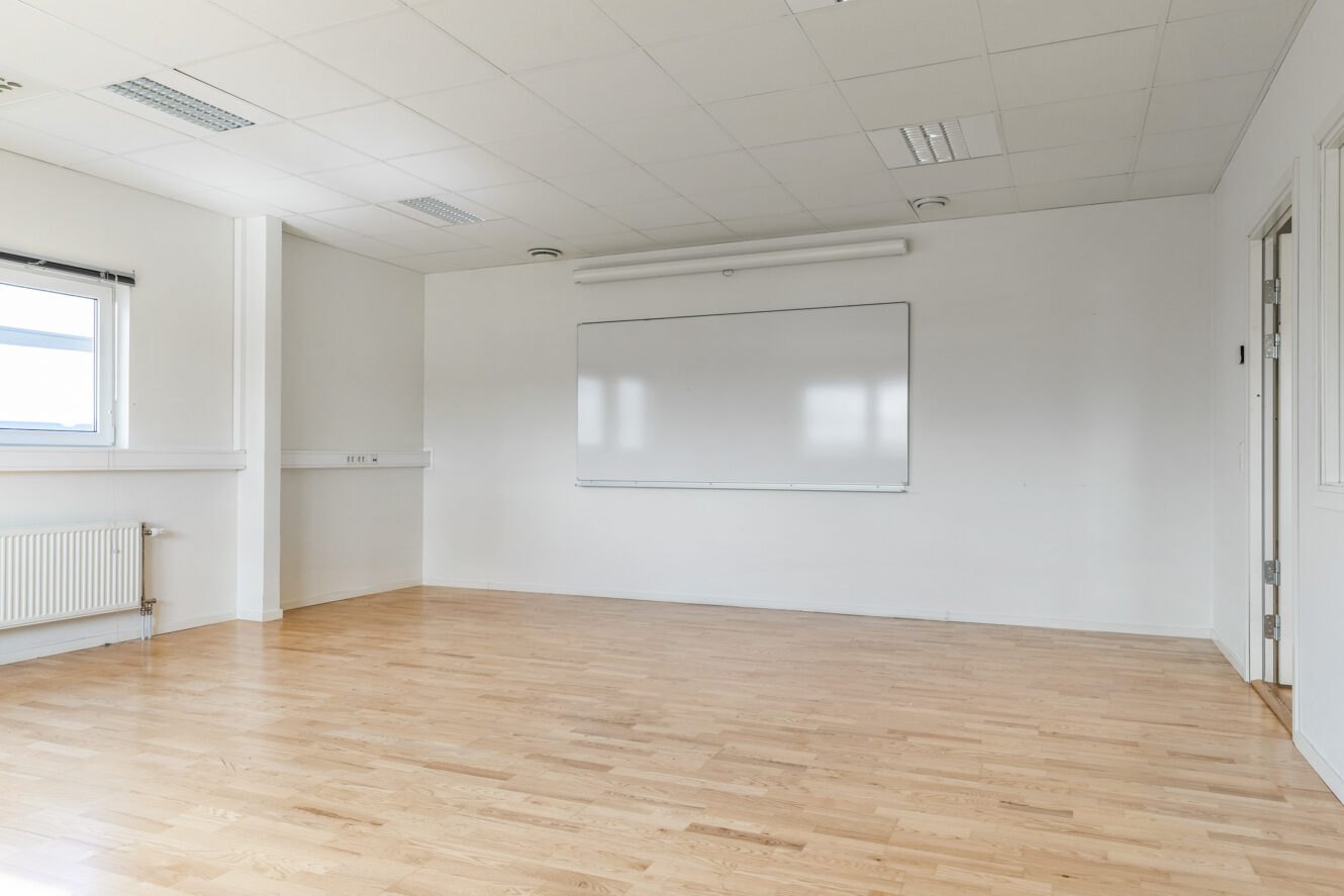 Ett tomt klassrum med ljust trägolv, vita väggar, en stor whiteboardtavla och ett fönster som släpper in dagsljus.