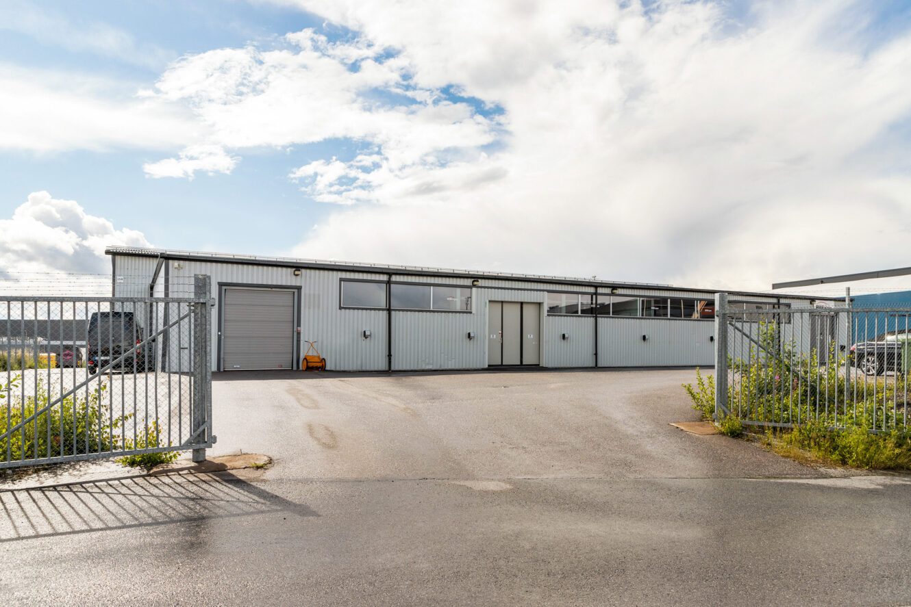 En industriell lagerlokal i metall med stora fönster och en stängd port framför, under en delvis molnig himmel.