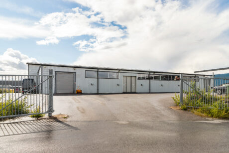 En industriell lagerlokal i metall med stora fönster och en stängd port framför, under en delvis molnig himmel.