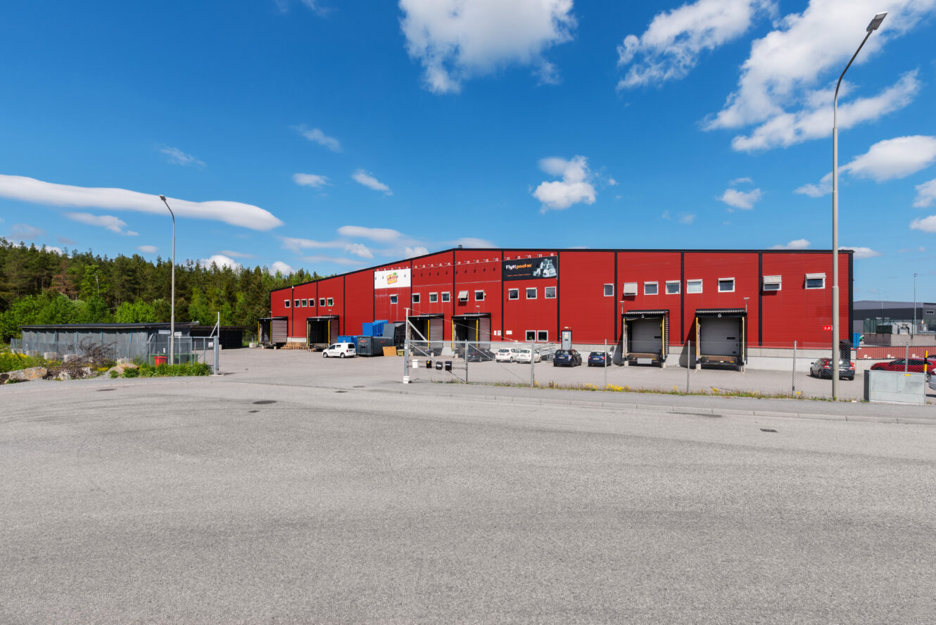 En stor röd lagerbyggnad med flera lastbryggor och parkerade bilar, omgiven av träd under en blå himmel med spridda moln.