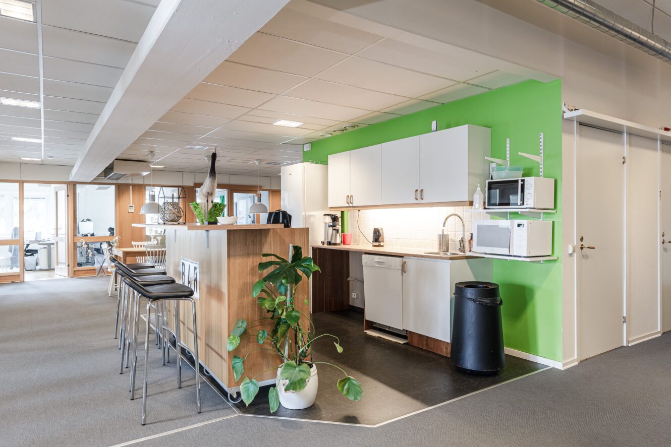 Modernt kontorskök med grön accentvägg, vita skåp, mikrovågsugnar, diskmaskin, barstolar, krukväxt och papperskorg; angränsar till korridor med stängda dörrar.