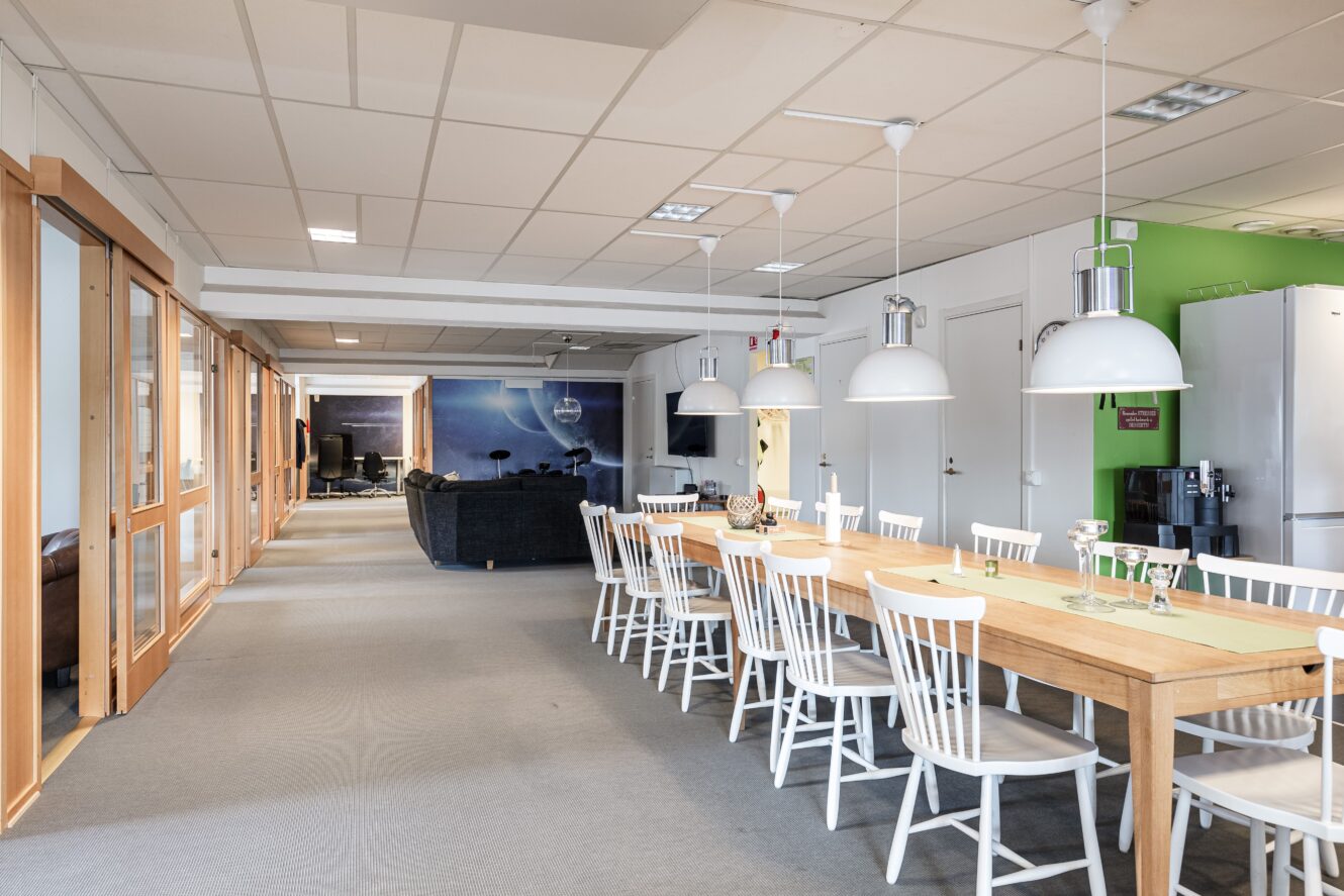 Ett modernt kontorskök och matplats med ett långt träbord, vita stolar, hängande lampor och en lounge i bakgrunden.