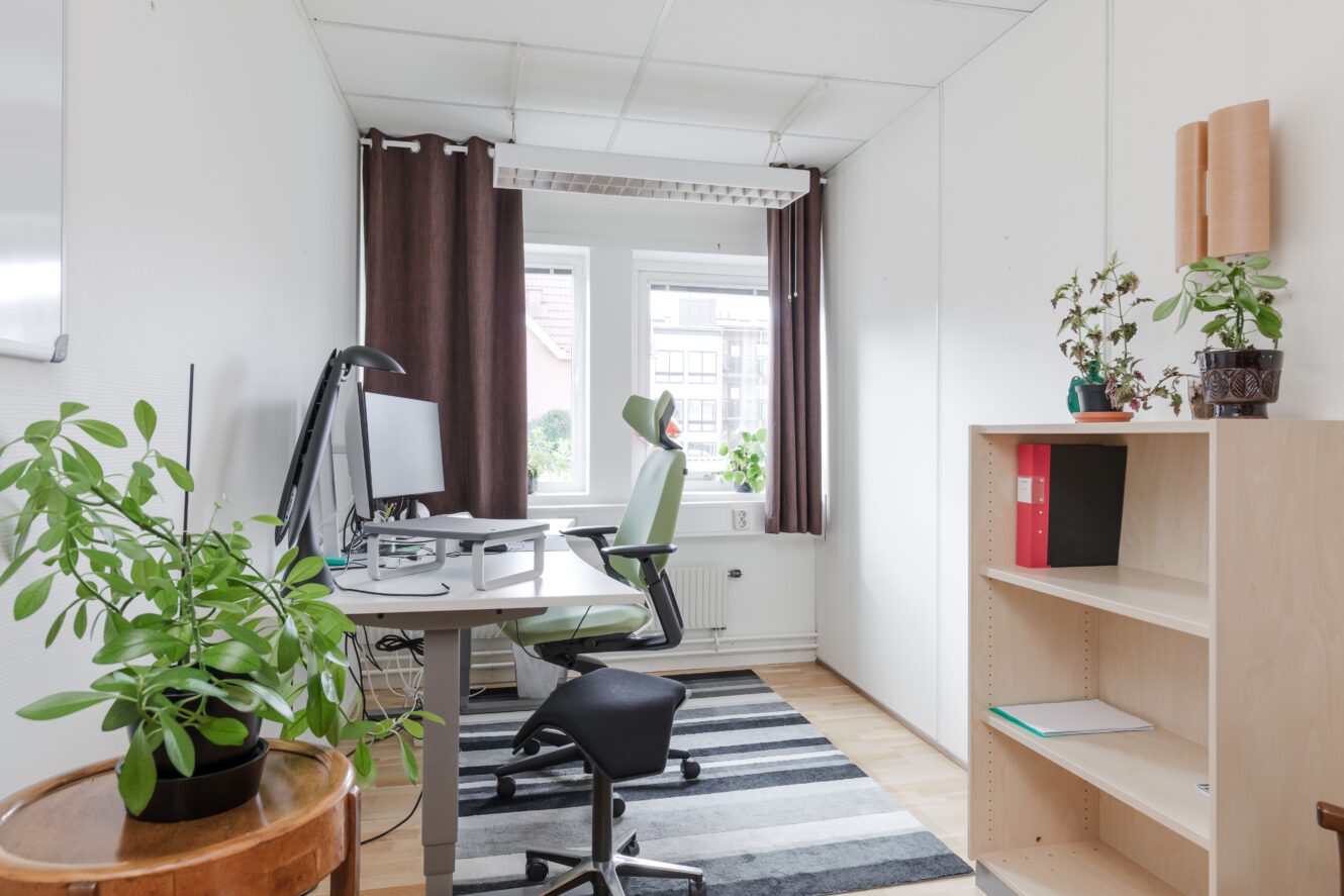 Modernt kontor med skrivbord, dator, kontorsstolar, växter, bokhylla med mappar, fönster med gardiner och en randig matta på ett ljust trägolv.