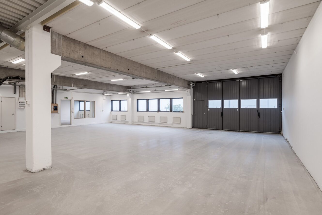 Rymlig, tom industrilokal med betonggolv, vita väggar, synliga bjälkar, lysrörsbelysning samt stora fönster och garageportar.