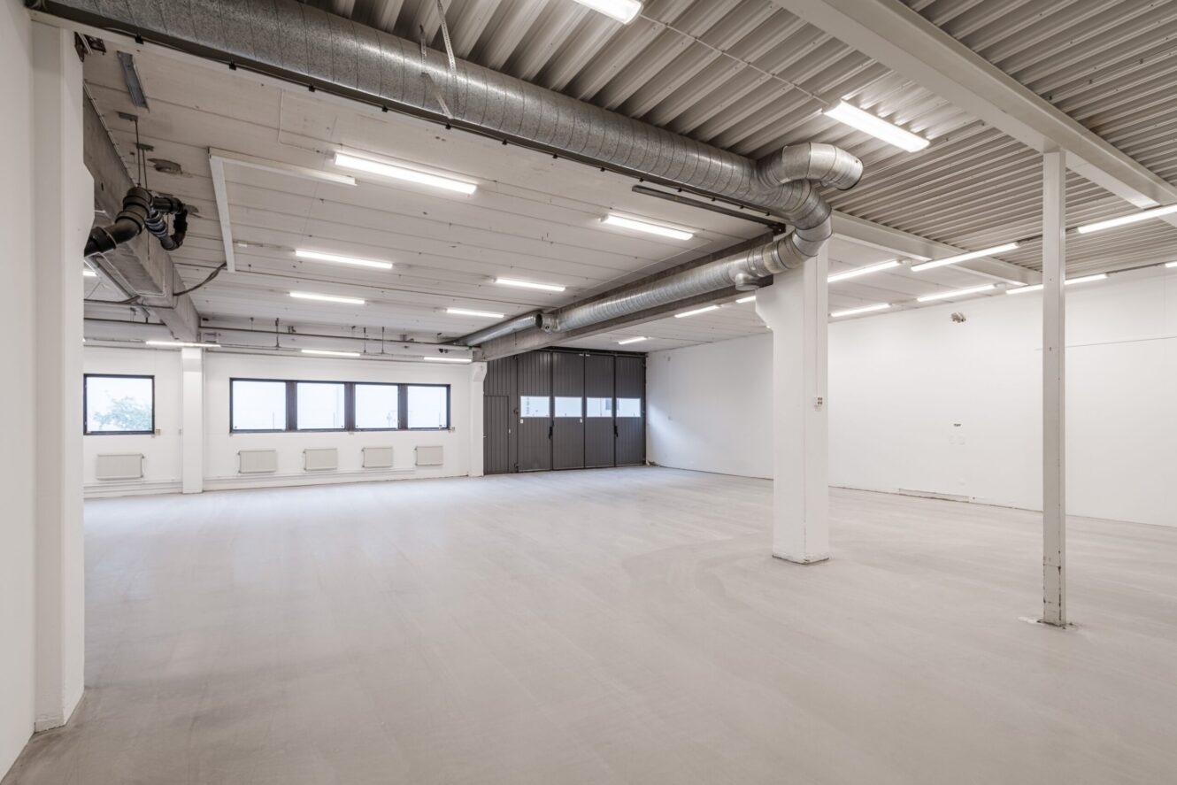 Rymlig, tom industrilokal med stora fönster, synliga takkanaler, vita väggar och betonggolv, upplyst av lysrör i taket.