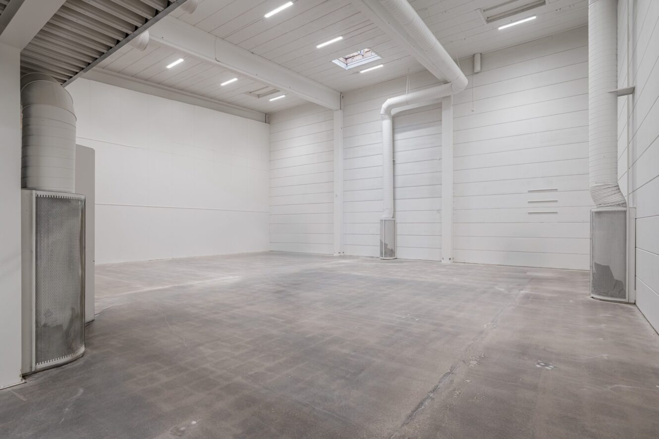 Tomt industriellt lagerutrymme med vita väggar, högt i tak, takbelysning, ventilationskanaler och polerat betonggolv.
