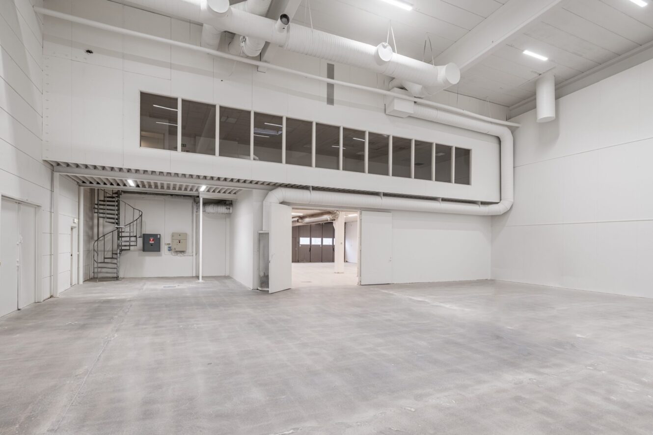 Tomt industrilager med betonggolv, vita väggar, högt i tak, synliga ventilationskanaler och ett slutet kontorsutrymme på övre plan med fönster och en metalltrappa.
