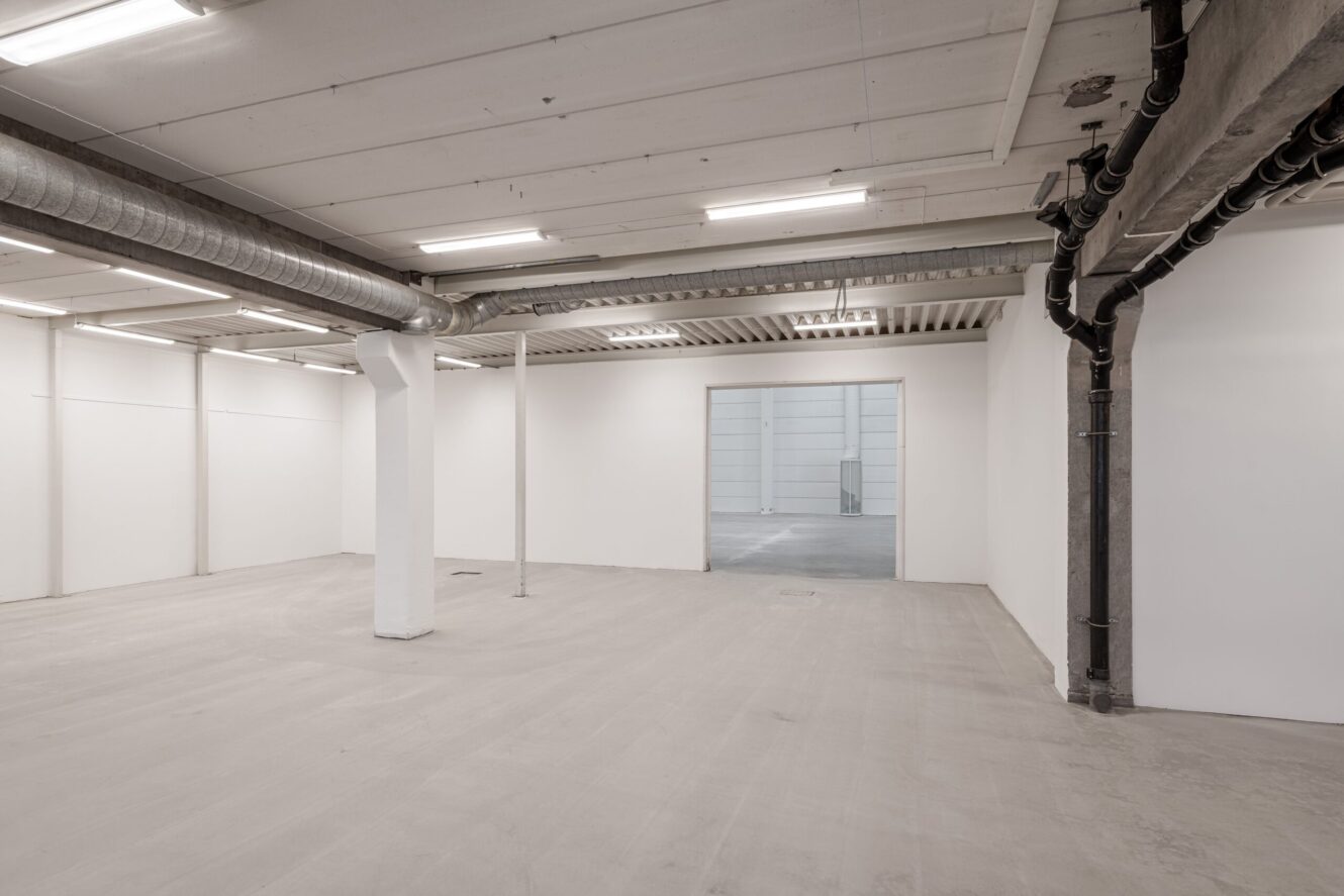 Tomt rum i industriell stil med vita väggar, synliga rör, lysrörsbelysning och en öppen dörr som leder till ett annat liknande utrymme.