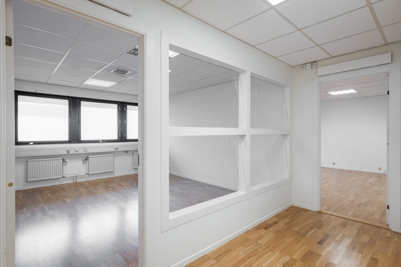 En modern, tom kontorslokal med vita väggar, stora fönster och trägolv. Ett rum är synligt genom glasväggen. I taket finns paneler med fluorescerande belysning.