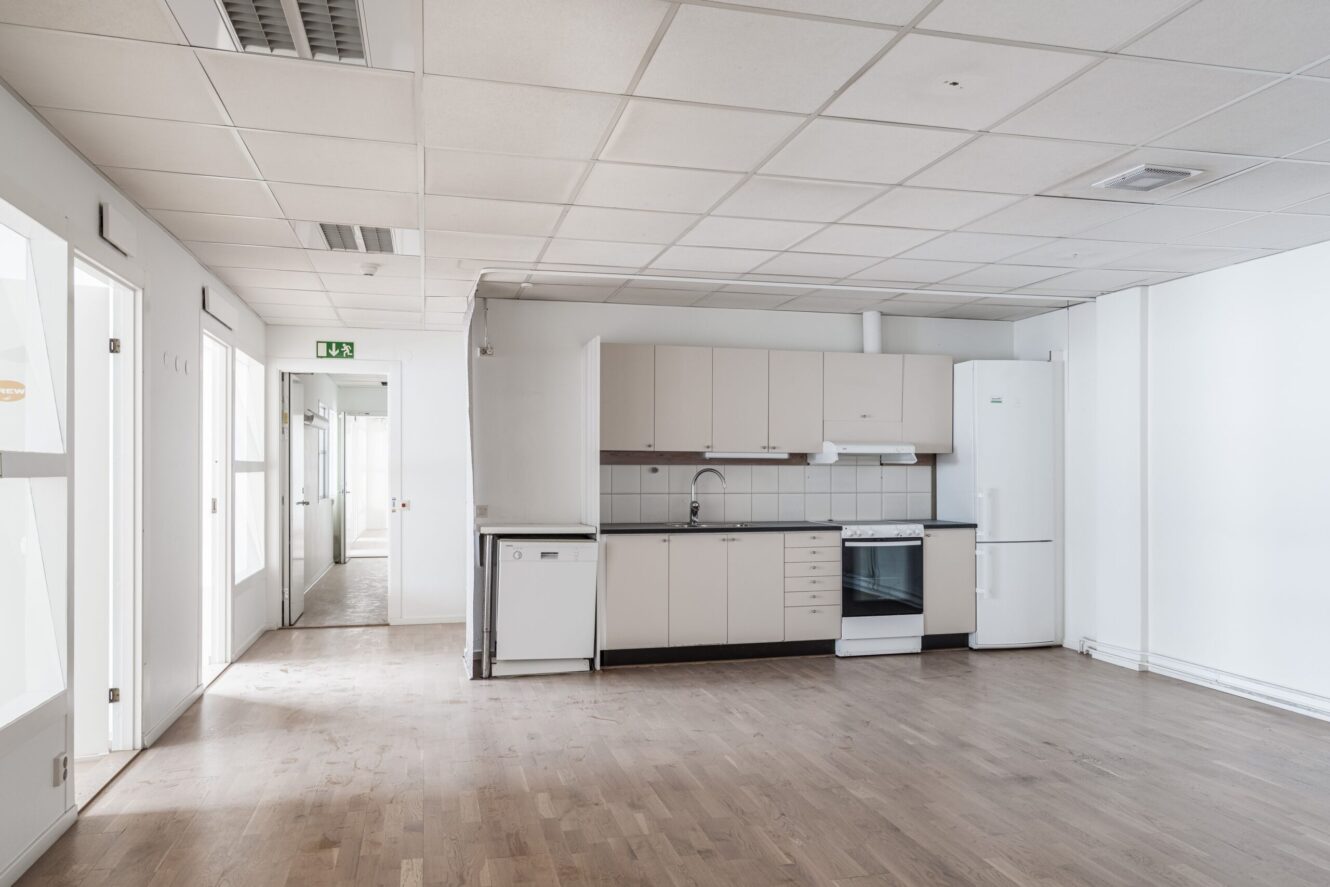 En tom, modern köksdel med ljusa skåp, vita vitvaror och trägolv, intill en hall i ett väl upplyst utrymme.