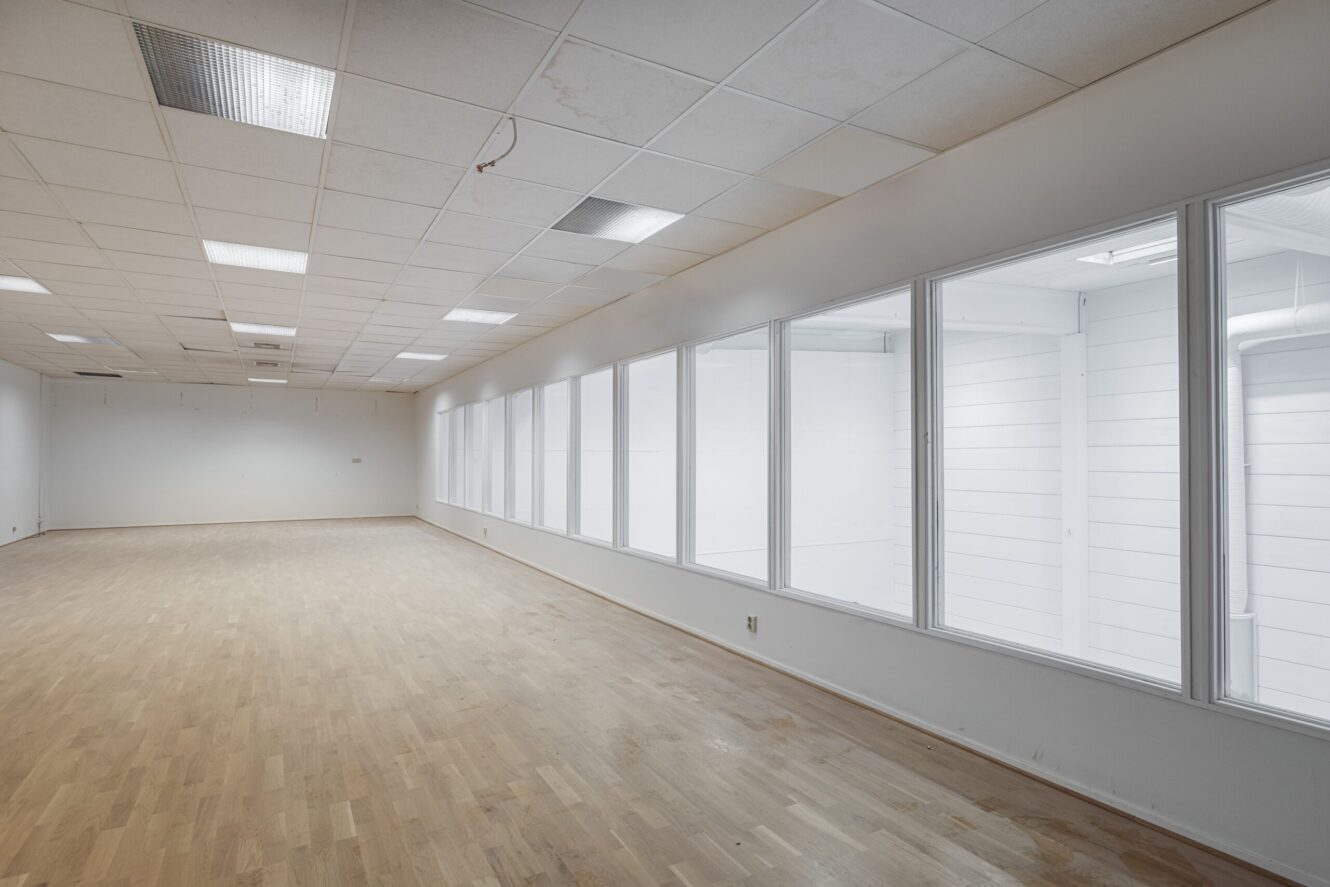 Tomt rum med ljust trägolv, vita väggar och en rad stora fönster på ena sidan, med taklampor och ett neddraget tak synligt.