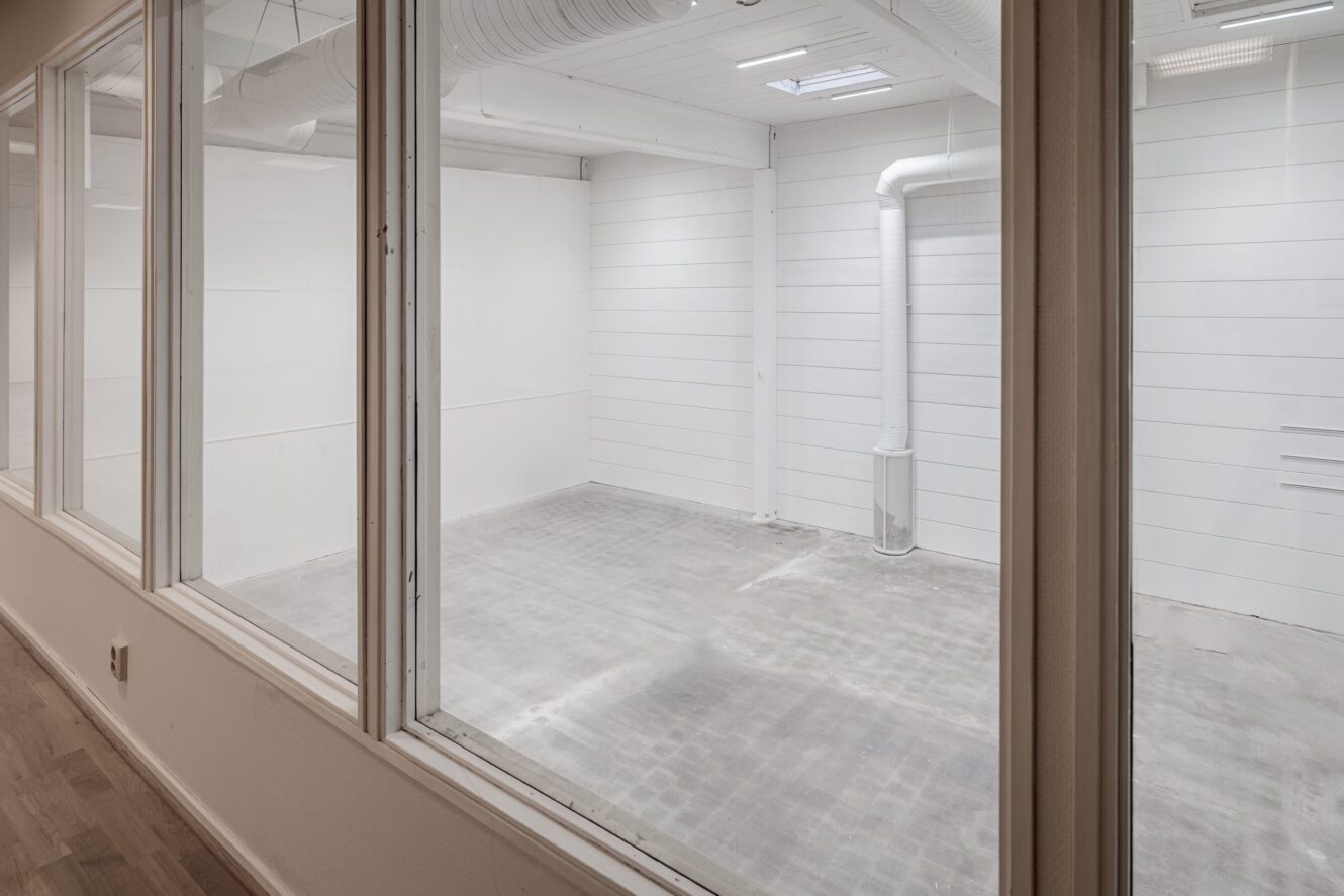 En vy genom glasfönster in i ett tomt industrirum med vita väggar, grått golv, synliga ventilationskanaler och synlig takbelysning.