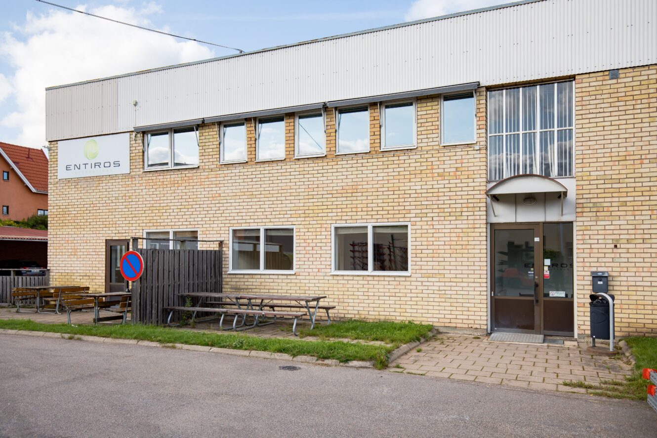 En tvåvånings tegelbyggnad med flera fönster, en ingångsdörr i glas, bänkar utanför och en skylt med texten "ENTIROS" på fasaden.