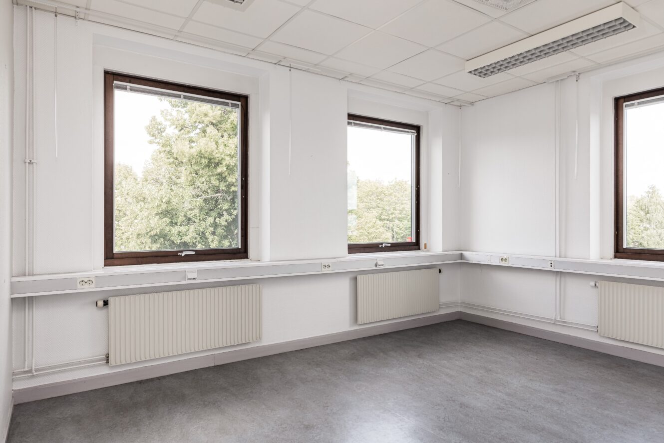 Tomt hörnkontor med två stora fönster, vita väggar, lysrör i taket och grått golv. Träd syns utanför genom fönstren.