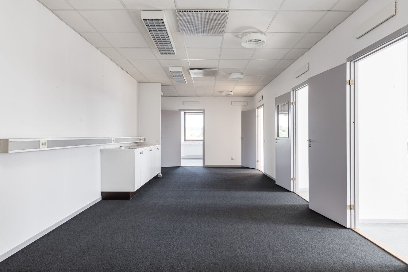 Tomt kontorsutrymme med vita väggar, mörk heltäckningsmatta, öppna dörrar, takbelysning och ett fönster längst bort.