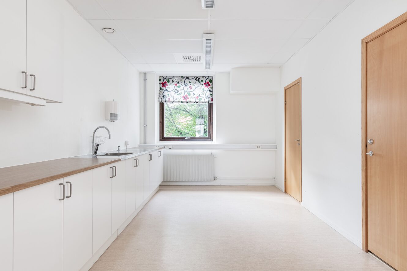 Ett minimalt kök med vita skåp, bänkskivor i trä, en diskho och ett fönster med en blommig gardin. Rummet är tomt och väl upplyst, med en stängd trädörr till höger.