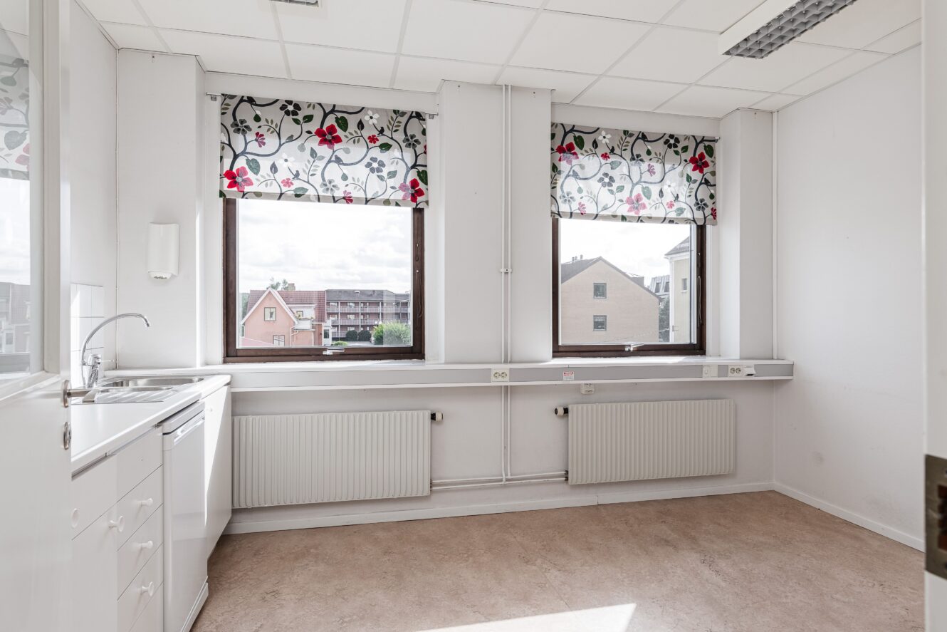 Ett ljust, tomt rum med två stora fönster, blommönstrade persienner, ett handfat, vita skåp och element under fönstren.