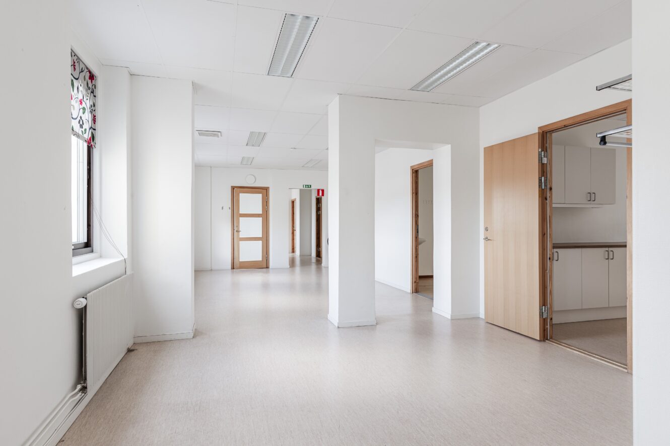 Ett ljust, tomt kontorsutrymme med vita väggar, beige golv, lysrör, flera trädörrar och ett fönster med en mönstrad gardin.