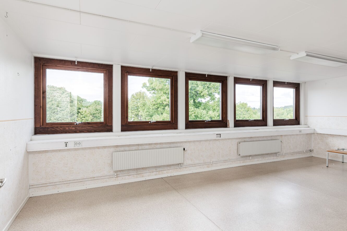 Tomt rum med beige golv, vita väggar och en rad med fem stora träfönster med utsikt över gröna träd. Två radiatorer är monterade under fönstren.