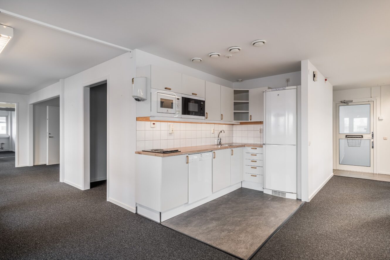 Modernt, kompakt vitt kök med inbyggda vitvaror, trädetaljer och kaklat stänkskydd i en öppen planlösning med grå matta och vita väggar.