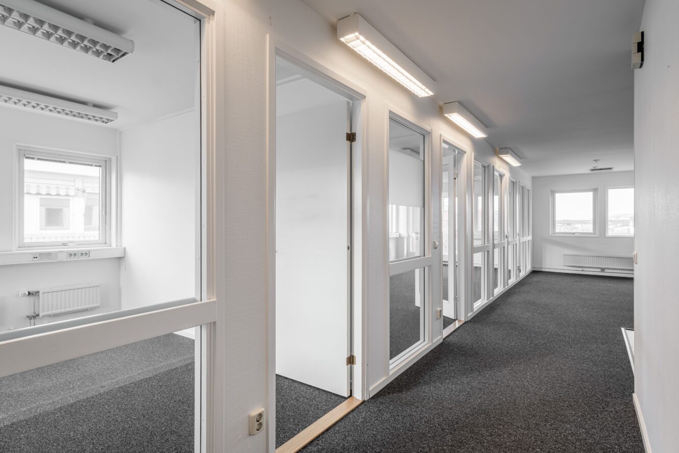 En korridor med heltäckningsmatta, vita väggar och flera kontorsdörrar med glaspaneler som leder till tomma rum med stora fönster.