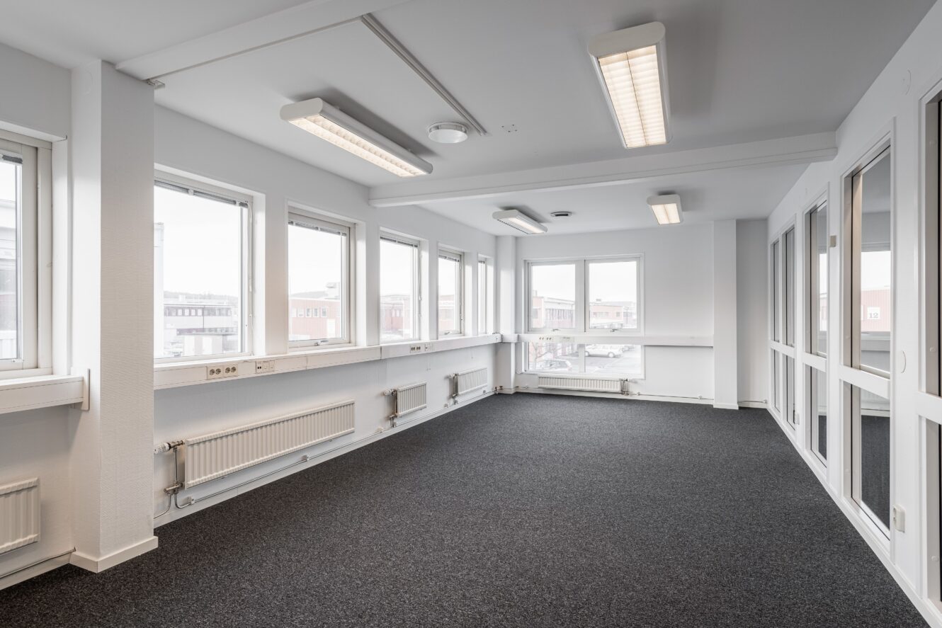 Ett ljust, tomt kontorsrum med stora fönster, vita väggar, grå heltäckningsmatta, taklampor och synliga radiatorer.