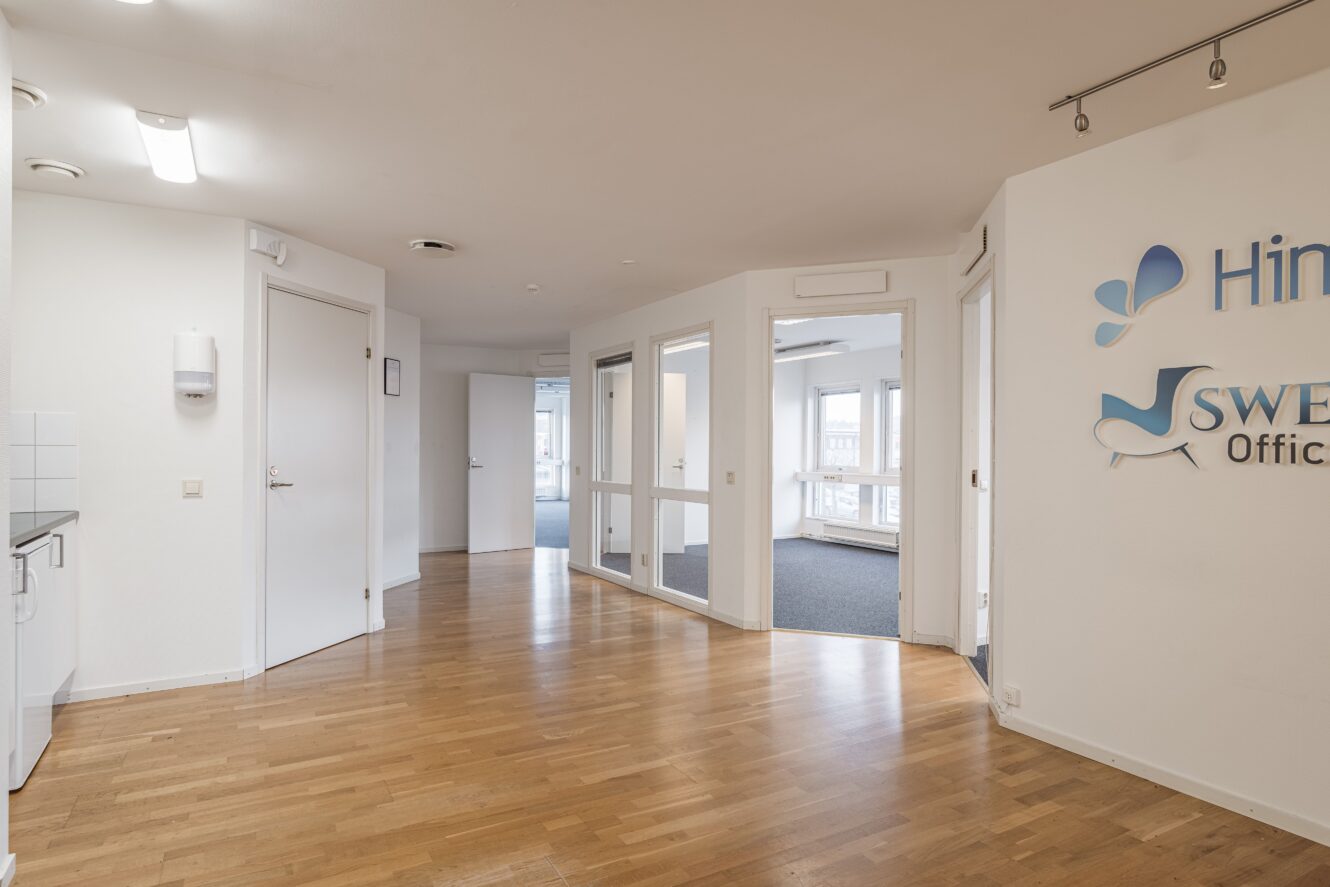 En modern kontorsinredning med ljusa väggar, trägolv, kontorsdörrar i glas och delvis skyltning på den högra väggen.