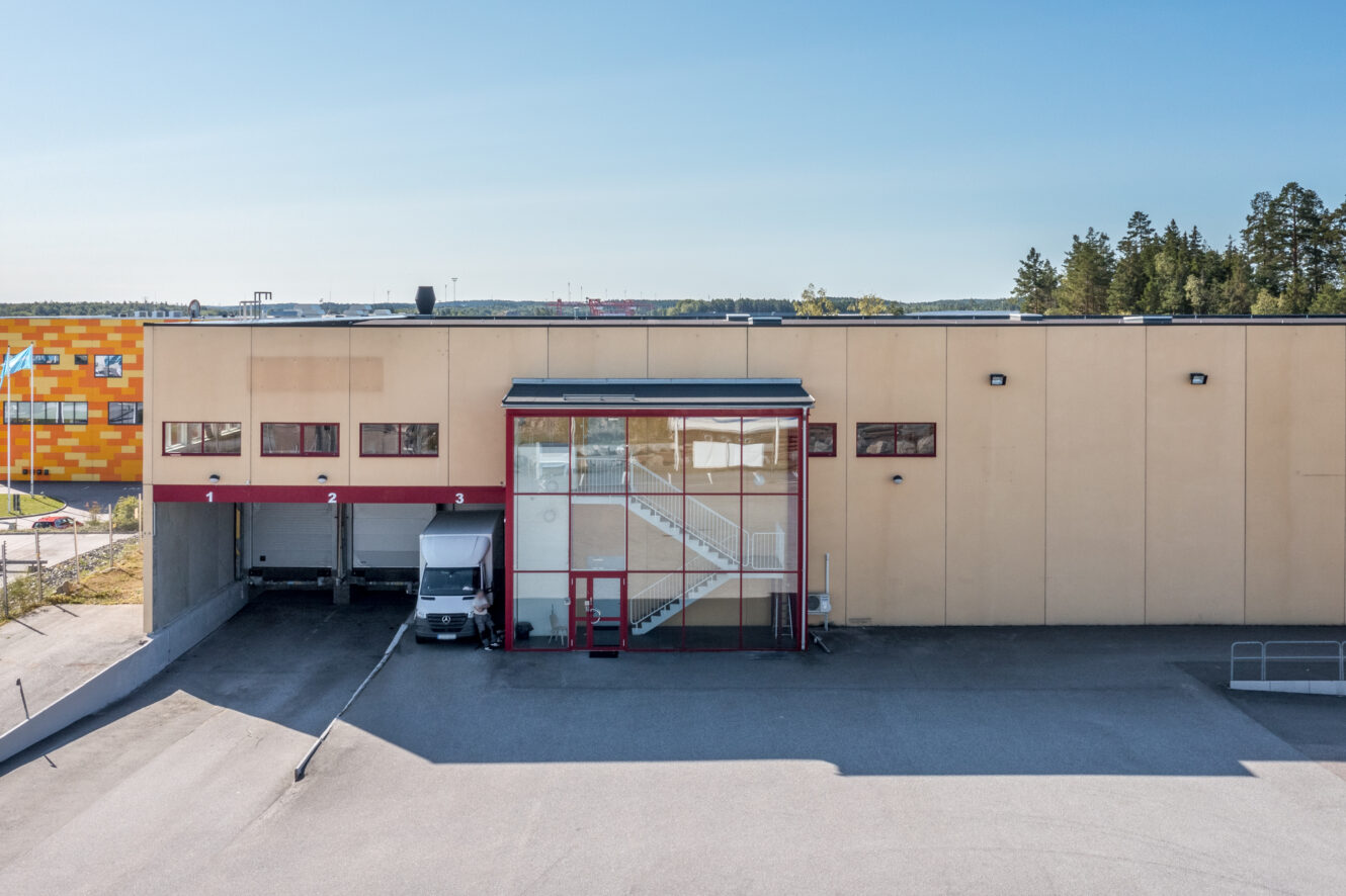 En beige industribyggnad med tre lastkajer, varav en upptagen av en vit lastbil, och en glasentré med synliga trappor, under en klar himmel.