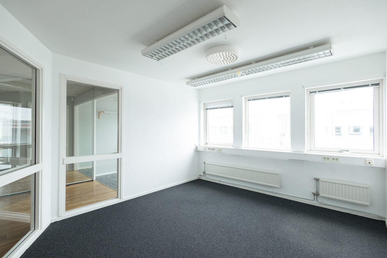 Tomt kontorsrum med vita väggar, grå heltäckningsmatta, tre stora fönster, lysrör i taket och en glasdörr som leder till ett annat rum.