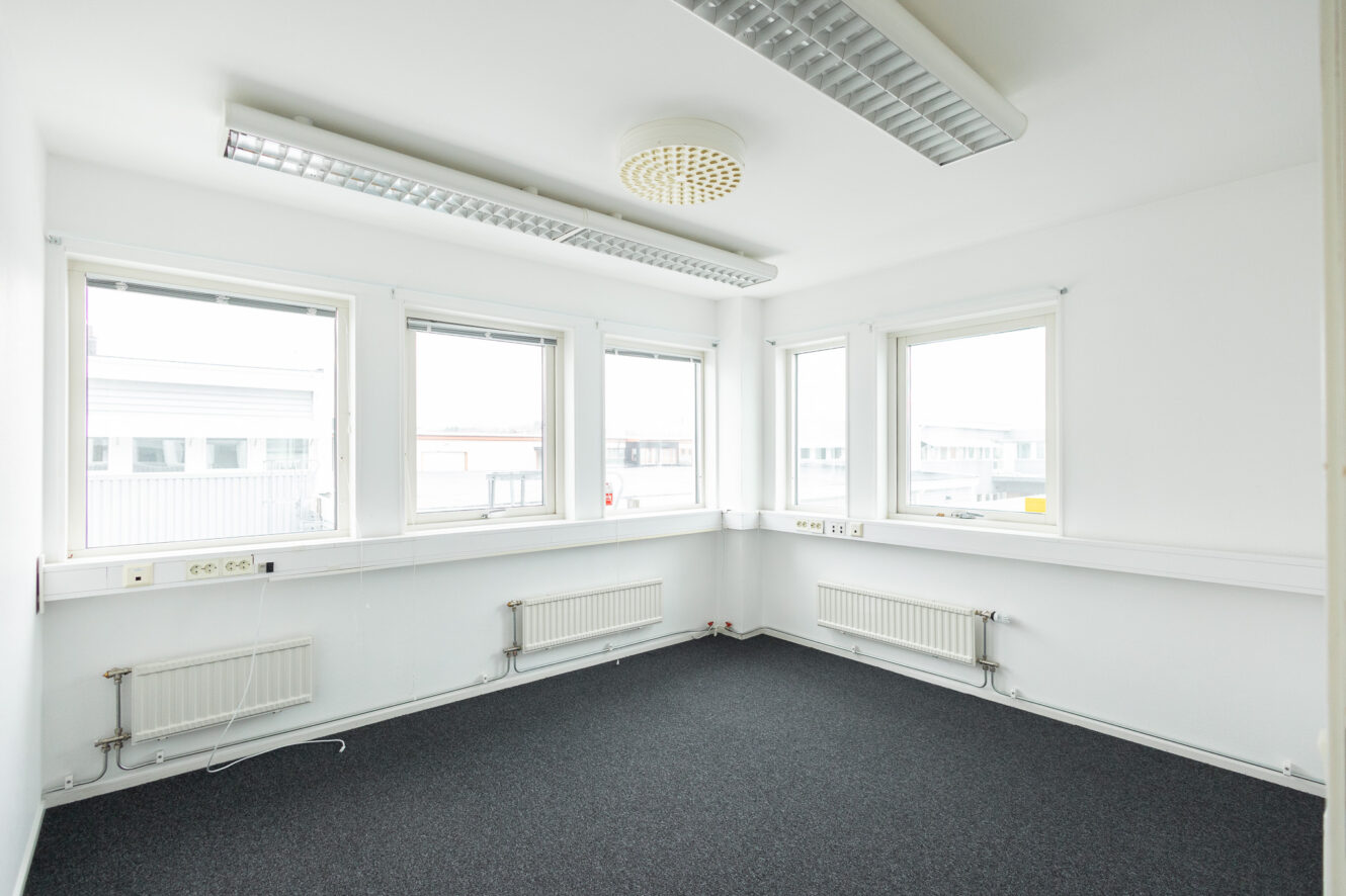 Ett tomt kontorsrum med vita väggar, tre stora fönster, grå heltäckningsmatta, två element och lysrör i taket.