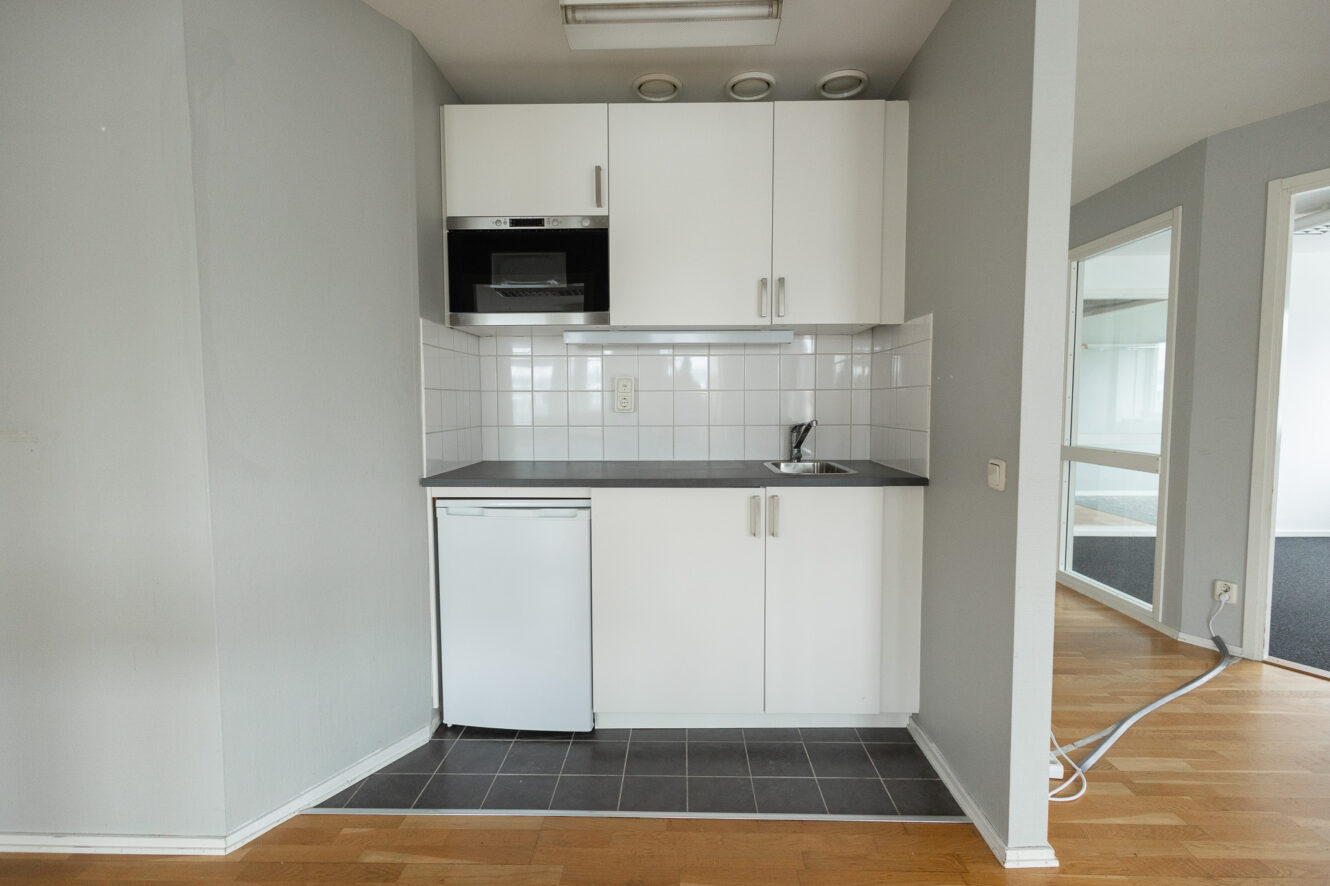 Kompakt pentry med vita skåp, ett litet kylskåp, diskho, spishäll, mikrovågsugn och kaklat stänkskydd mot en grå vägg; trä- och klinkergolv synligt.