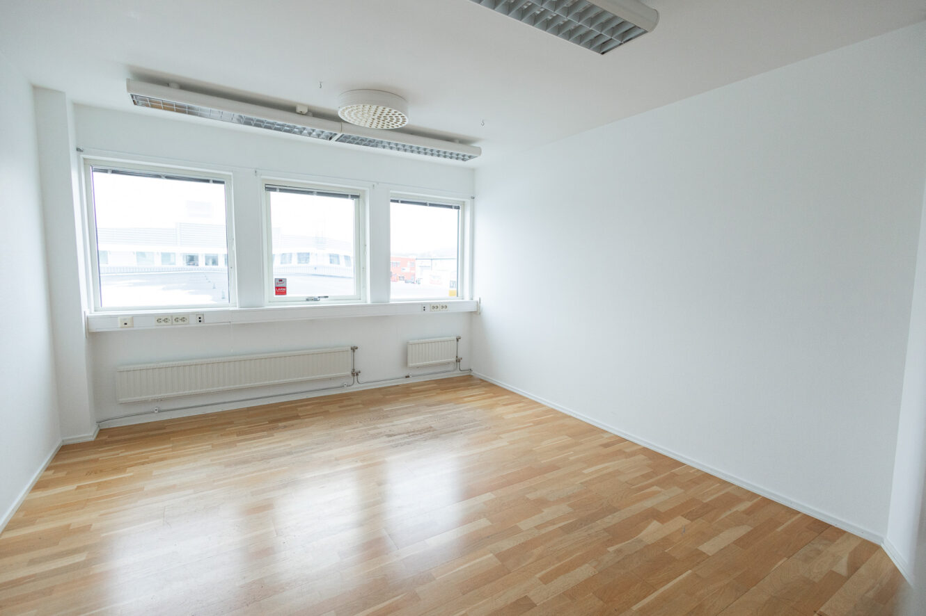 Tomt vitt rum med tre fönster, ljust trägolv, väggvärmare och taklampor.