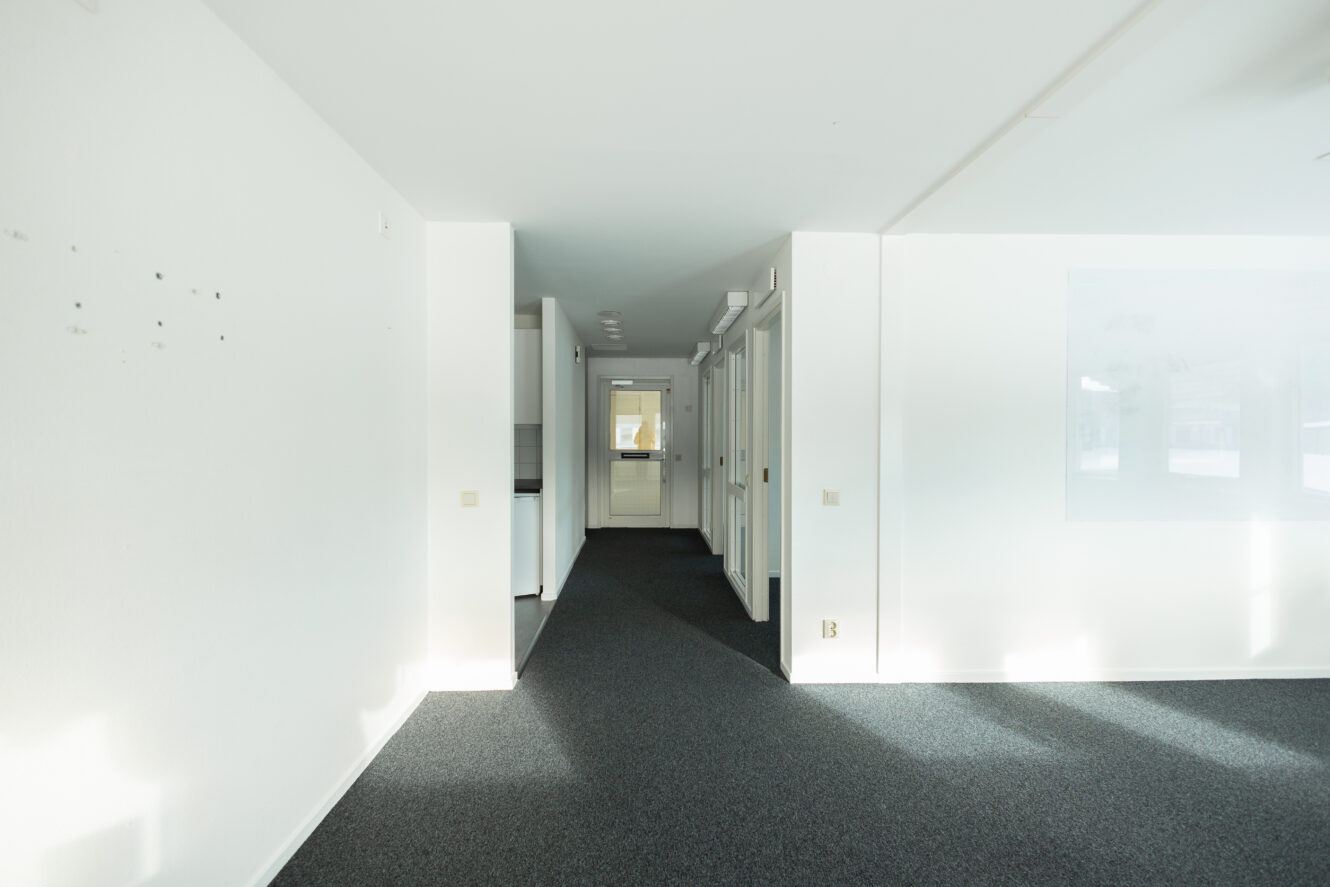 En korridor i en tom kontorsbyggnad med vita väggar, flera dörrar till höger och solljus som strömmar in genom fönstren.