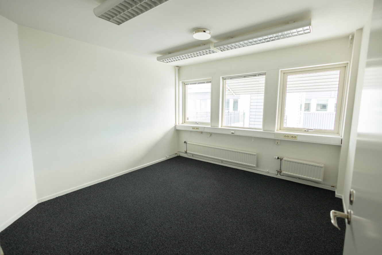 Tomt kontorsrum med vita väggar, mörk heltäckningsmatta, tre fönster med persienner, lysrör i taket och synliga radiatorer under fönstren.