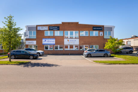 En tvåvånings affärsbyggnad med flera företagsskyltar på fasaden, parkerade bilar framför och en klarblå himmel ovanför.