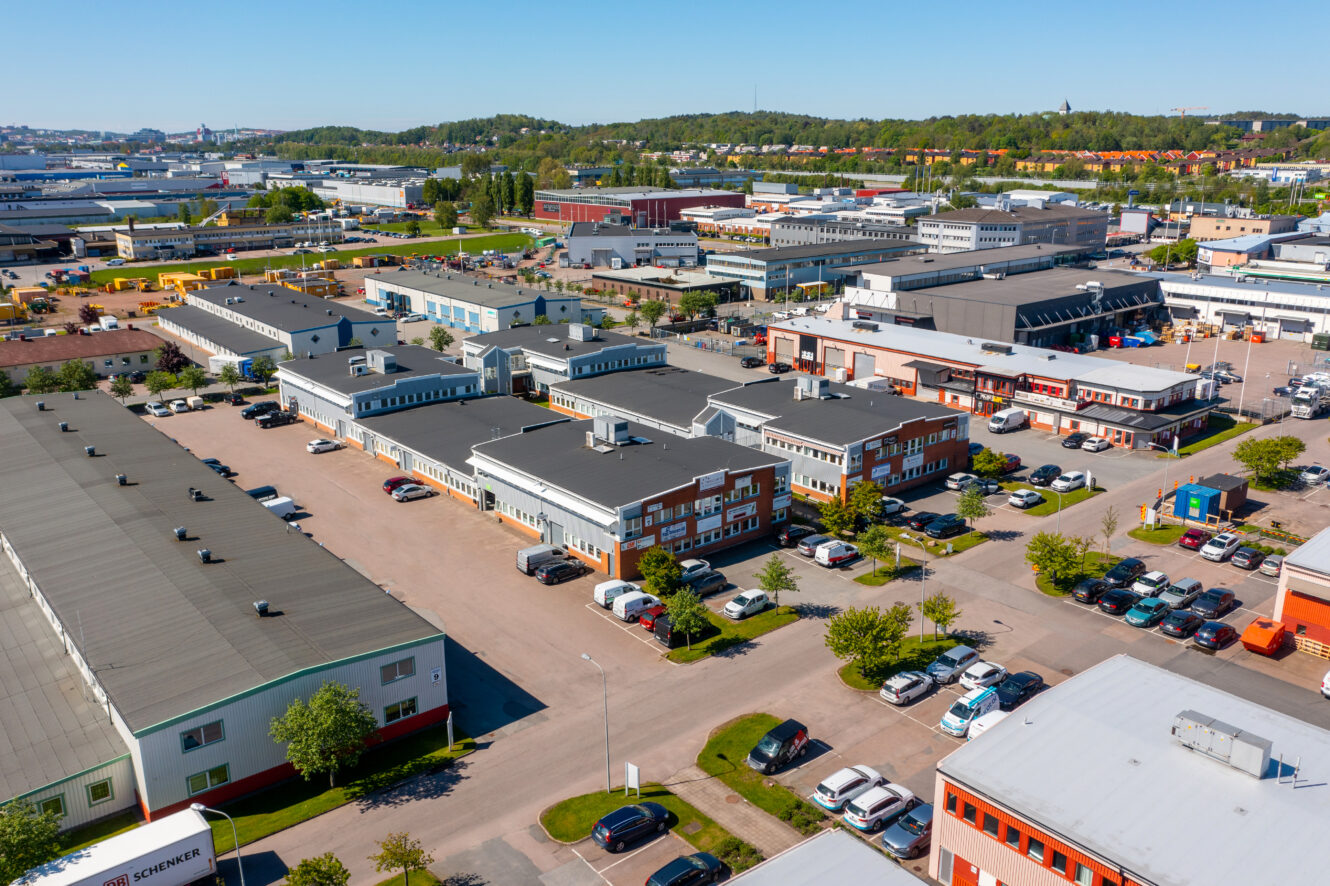 Flygfoto över en industripark med flera lagerlokaler, kontorsbyggnader, parkerade bilar och omgivande grönska under en klar himmel.