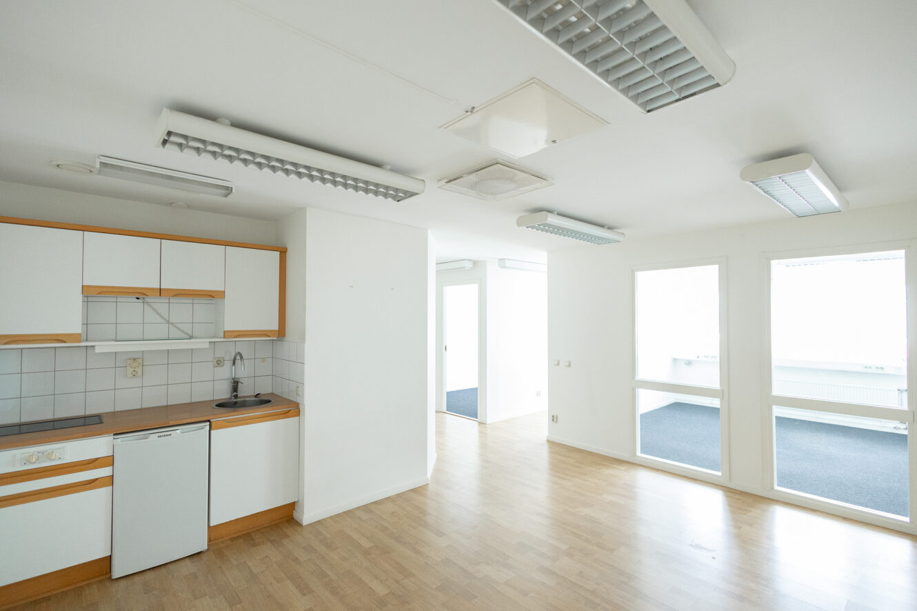 Ett litet kök med vita skåp och träinredning öppnar sig mot ett ljust, tomt rum med ljust trägolv och stora fönster. Lysrör är monterade i taket.