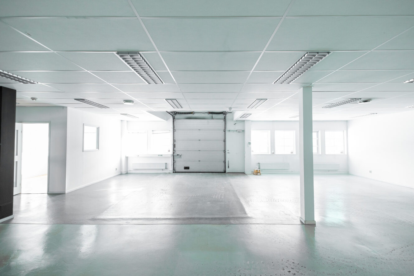 Ljus, tom industrilokal med vita väggar, lysrör i taket, stora fönster och en garageport på baksidan.