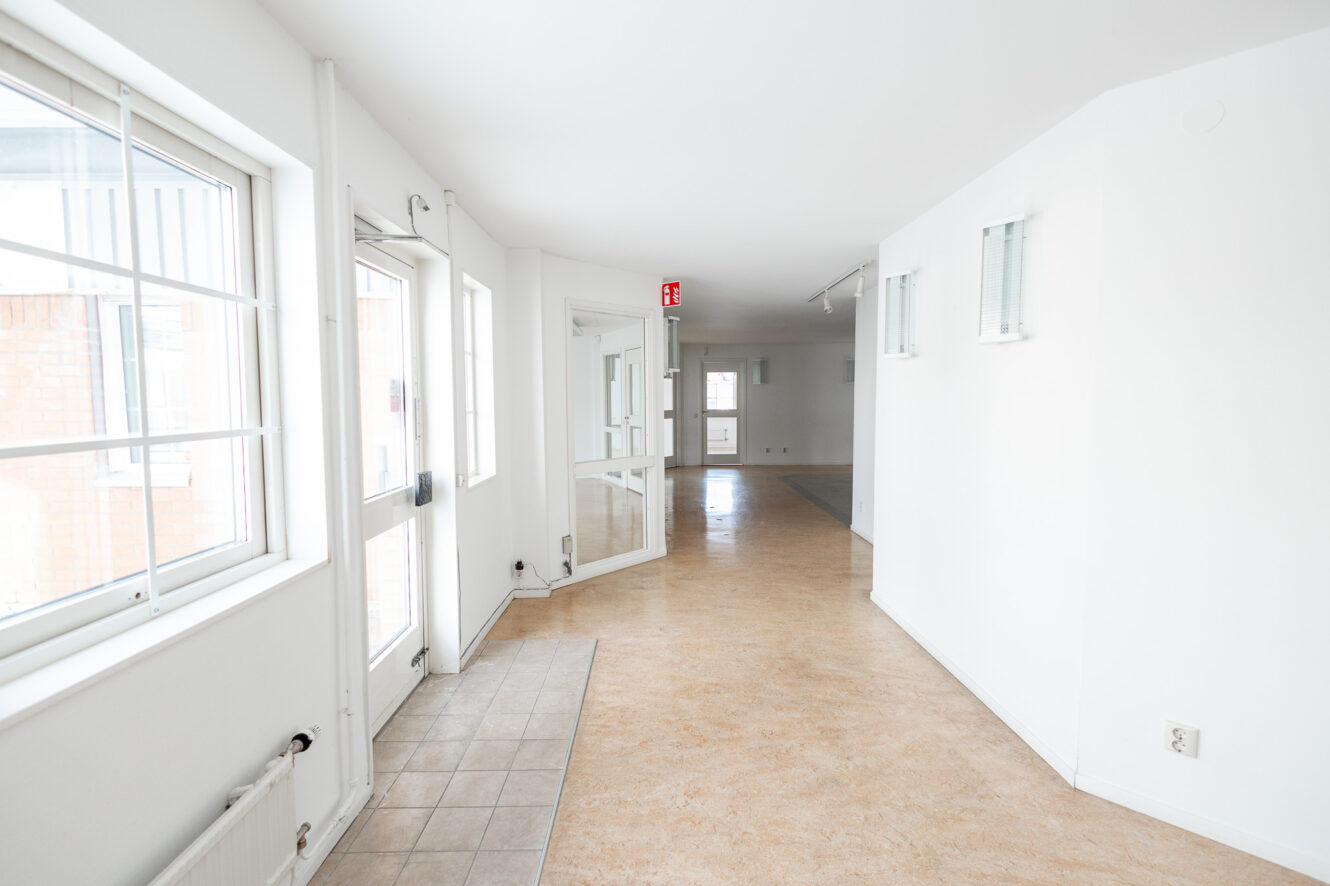 Ljus, tom hall med vita väggar, beige golv, stora fönster, en glasdörr och en nödutgångsskylt i taket.