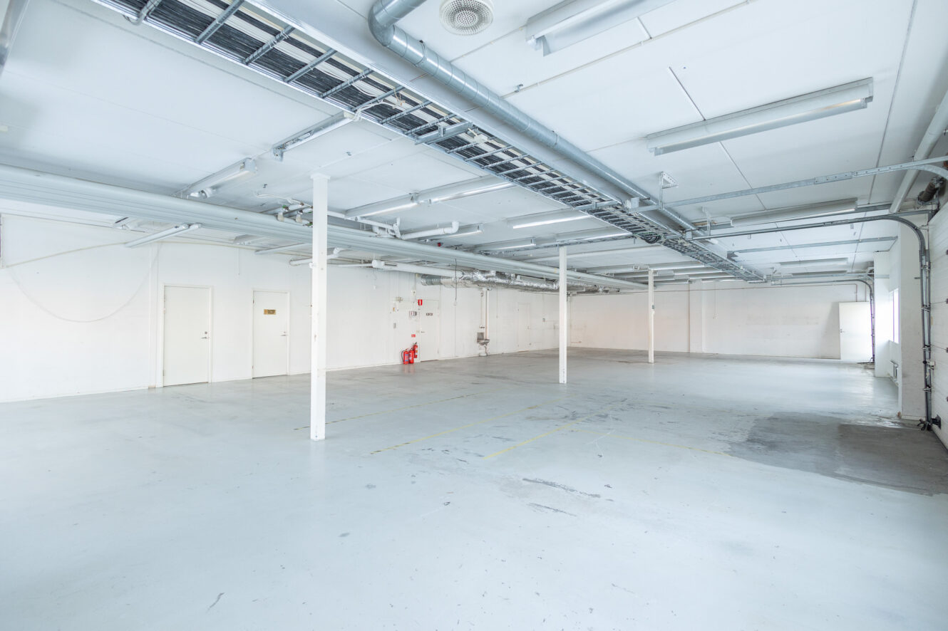 Stort, tomt industrilager med vita väggar, synliga rör i taket, stödpelare, brandsläckare och ett betonggolv med svaga linjer.