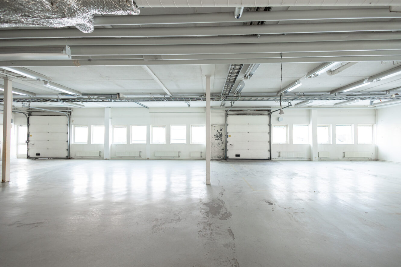 Tomt industrilager med vita väggar, stora fönster och rörledningar. Två garageportar syns på baksidan och betonggolvet visar tecken på slitage.