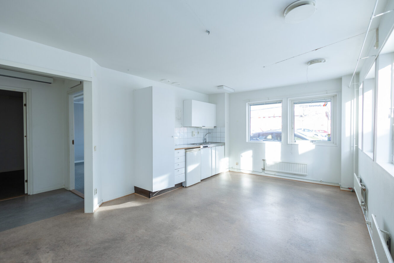 Omöblerad, ljus studiolägenhet med ett litet pentry, stora fönster, vita väggar och en öppen planlösning.