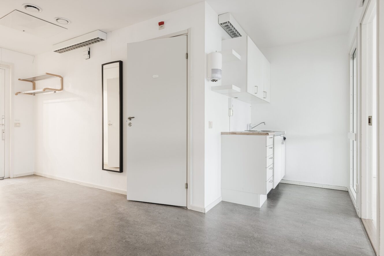 Ett litet, tomt rum med en stängd dörr, väggmonterad spegel, klädhängare och ett kompakt pentry med skåp och diskho. Golvet är grått och väggarna är vita.