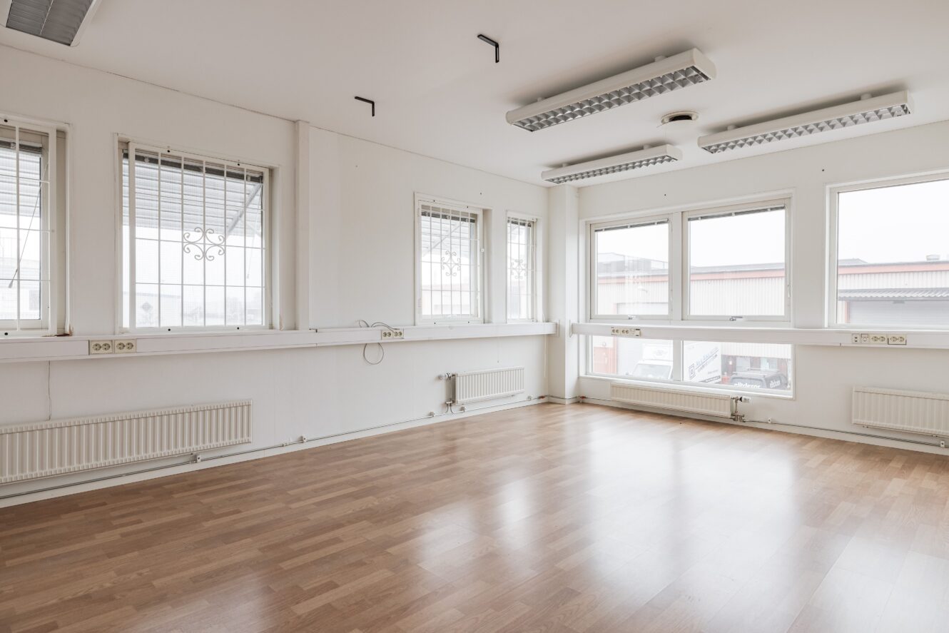Tomt rum med ljusa väggar, stora fönster med säkerhetsgaller, trägolv, lysrör i taket och synliga radiatorer och eluttag längs väggarna.
