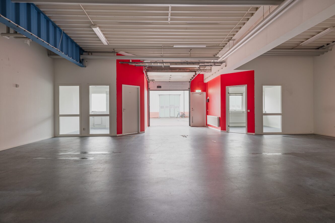 Tom industrilokal med grått betonggolv, vita och röda väggar och flera små rum med glasdörrar och fönster; i taket finns synliga rör och bjälkar.
