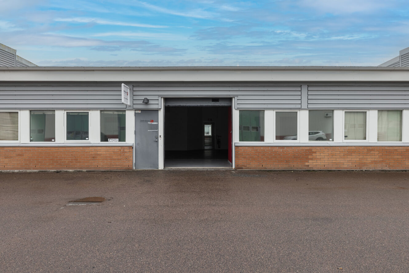 En envånings affärsbyggnad med fasad av tegel och grå panel, vitkarmade fönster och en öppen garageport som vetter mot en våt asfaltsyta.