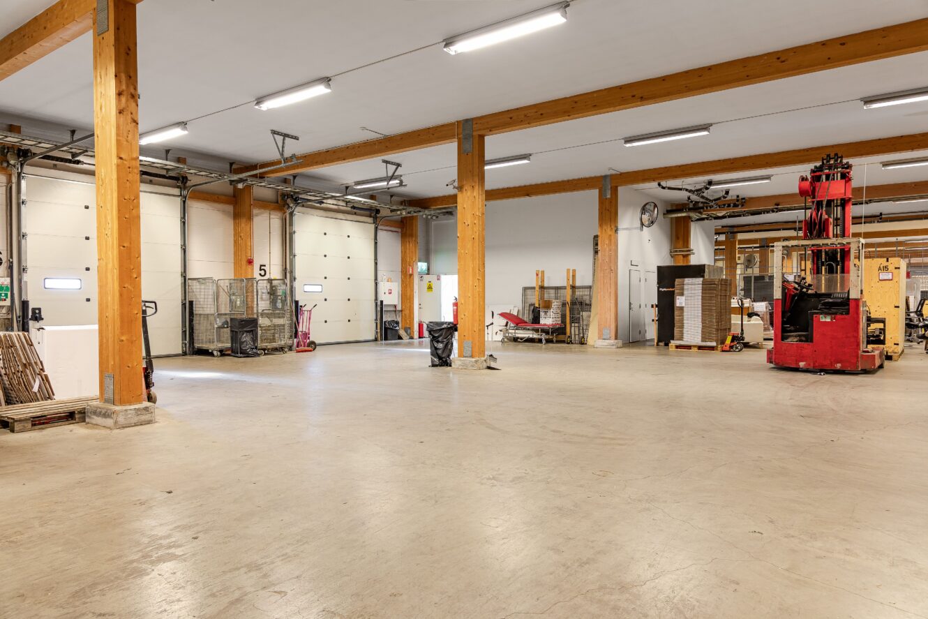 Rymlig lagerlokal med betonggolv, stödbalkar av trä, en röd gaffeltruck, lagerhyllor, pallar och diverse utrustning.