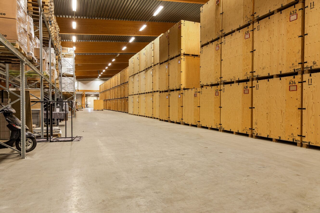 En lagerlokal med höga staplar av stora trälådor längs den högra väggen och metallhyllor med olika föremål till vänster. Golvet är rent och området är väl upplyst.